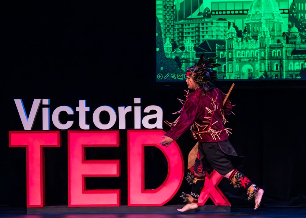 An Indigenous dancer does a dramatic leap in front of glowing red TEDx Victoria letters and green backdrop at Victoria TED event.