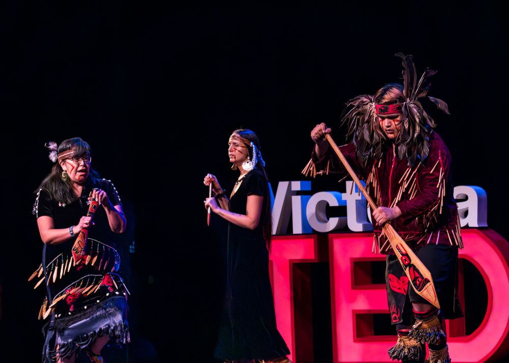 Three Indigenous performers on stage at TEDx Victoria with colourful lighting and dramatic poses during a live performance.