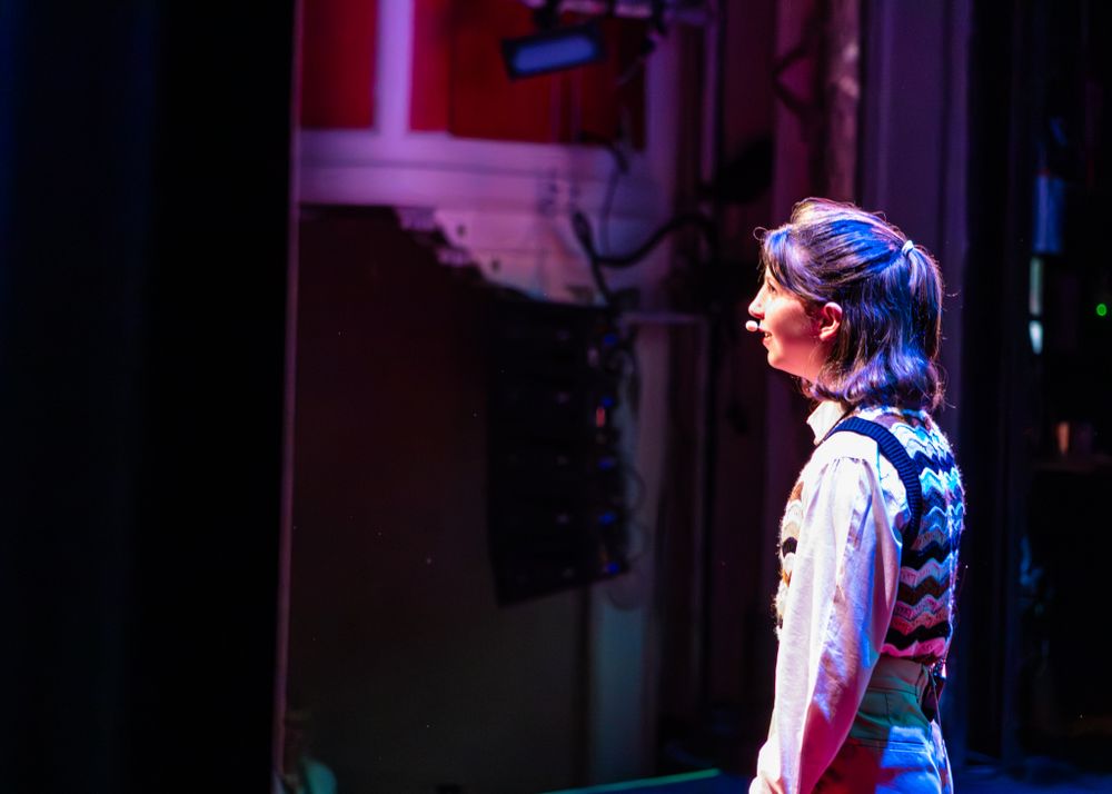 A performer stands in profile on stage wearing a traditional braided hairstyle and white shirt under blue theatrical lighting.