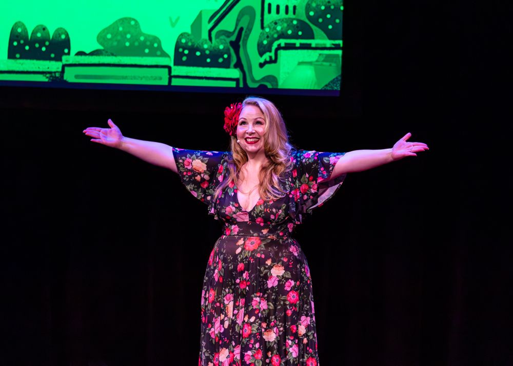 A woman in a floral dress stands on stage with arms outstretched against a dark background.