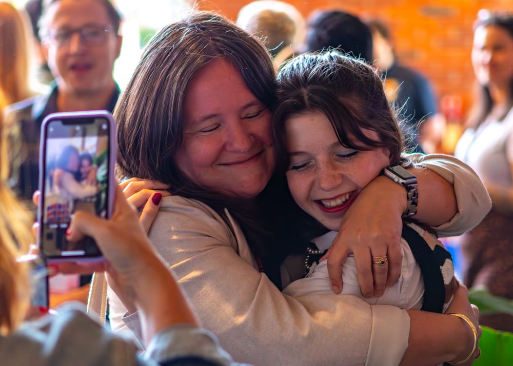 Two people share a joyful embrace while someone captures the moment on a phone in the background.