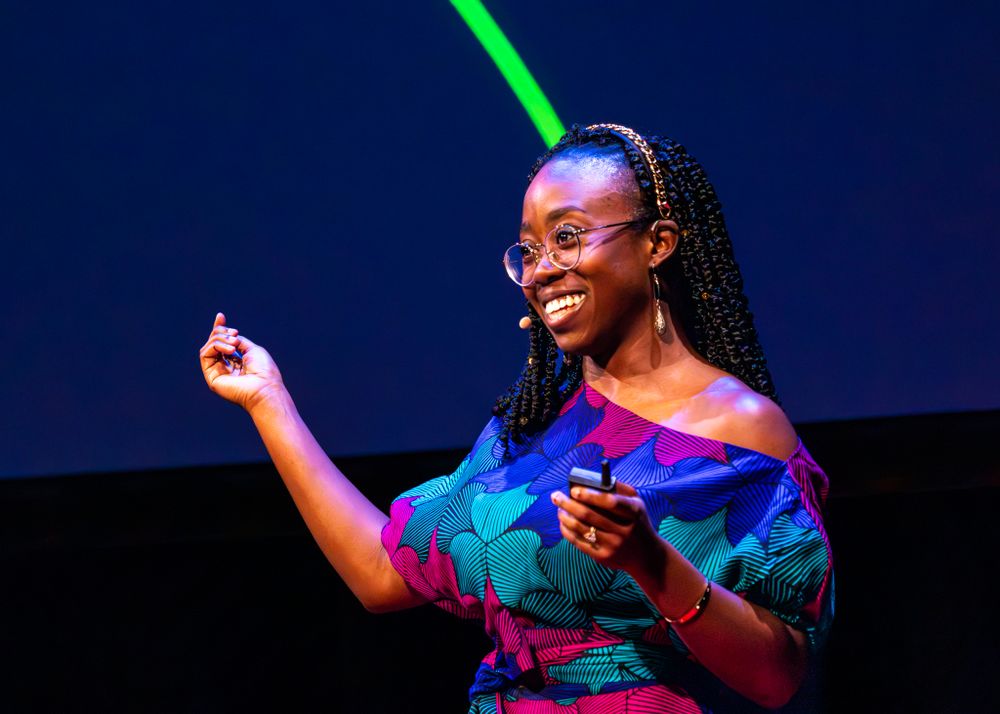 Speaker on stage wearing colorful teal and purple dress gestures while smiling under blue stage lighting.