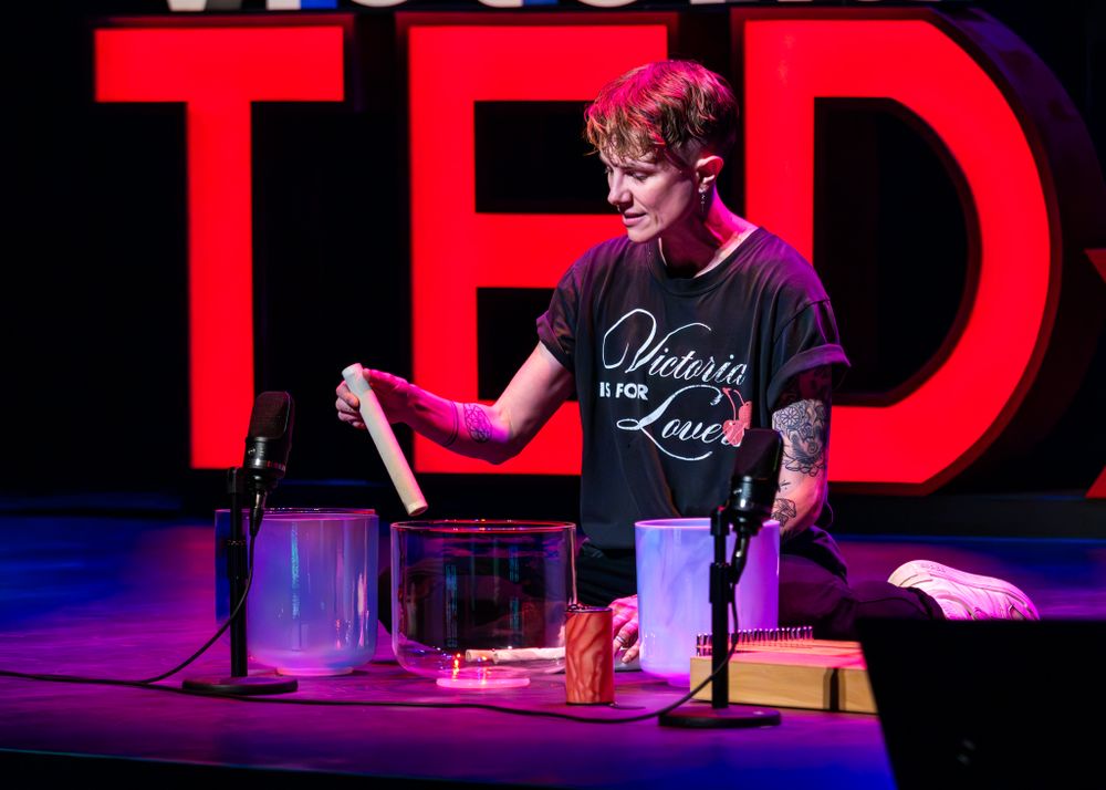 Speaker stands on stage at TED conference with large red TED logo illuminated behind them in dark theater setting.