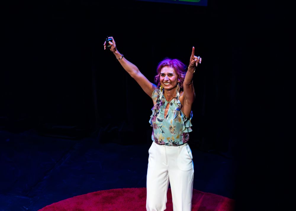 Speaker stands triumphantly with arms raised on circular red stage at TED talk event with blue lighting.