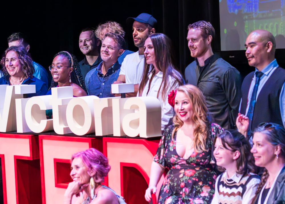 A group of performers stand together on stage behind large red and white TEDx letters during a presentation.