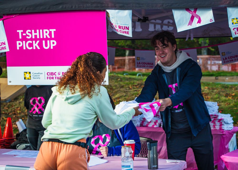 Volunteers distribute participant shirts at an outdoor event station with pink signage and boxes.