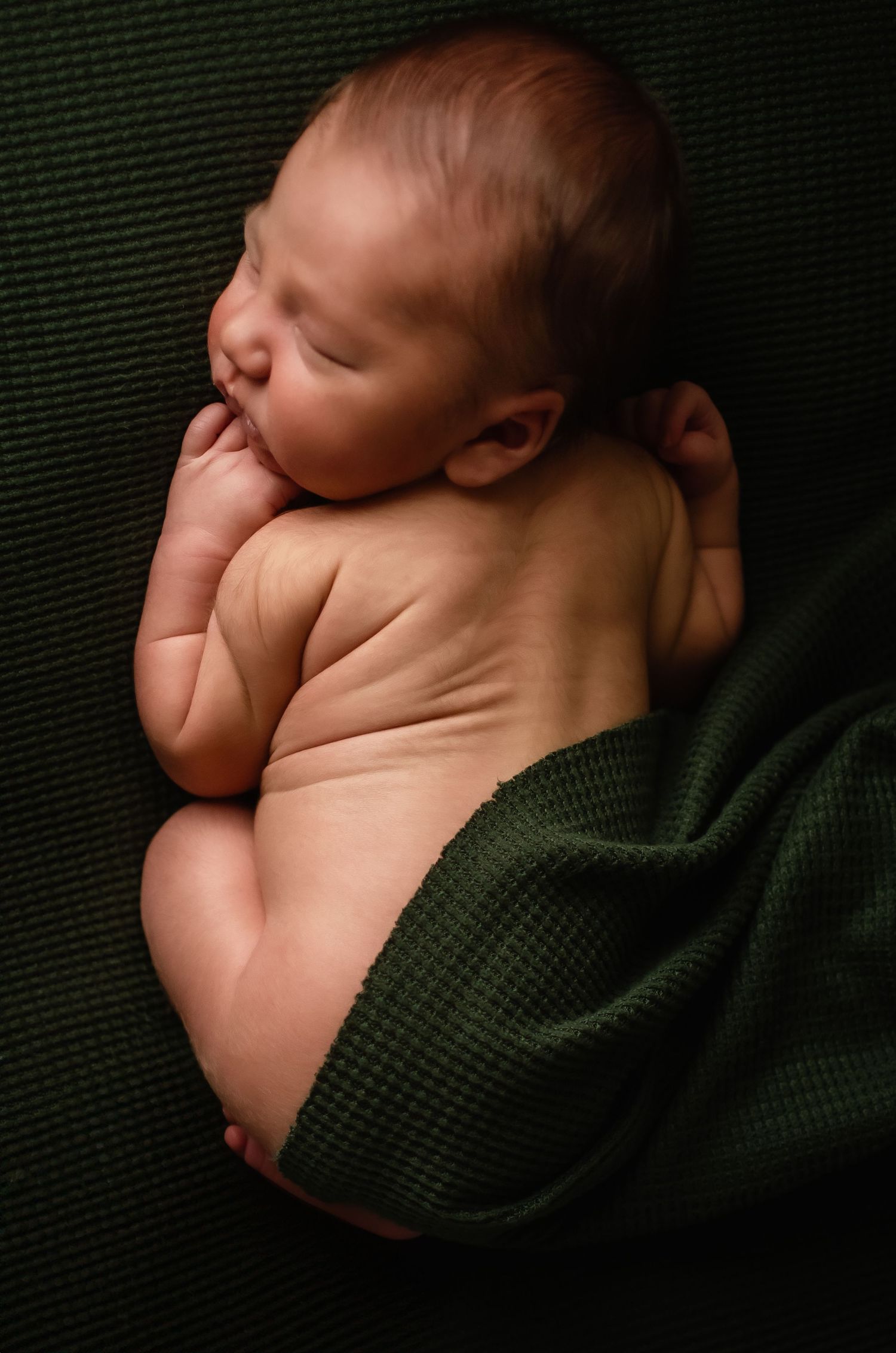 Peaceful newborn baby sleeping on dark green knit blanket in soft lighting.