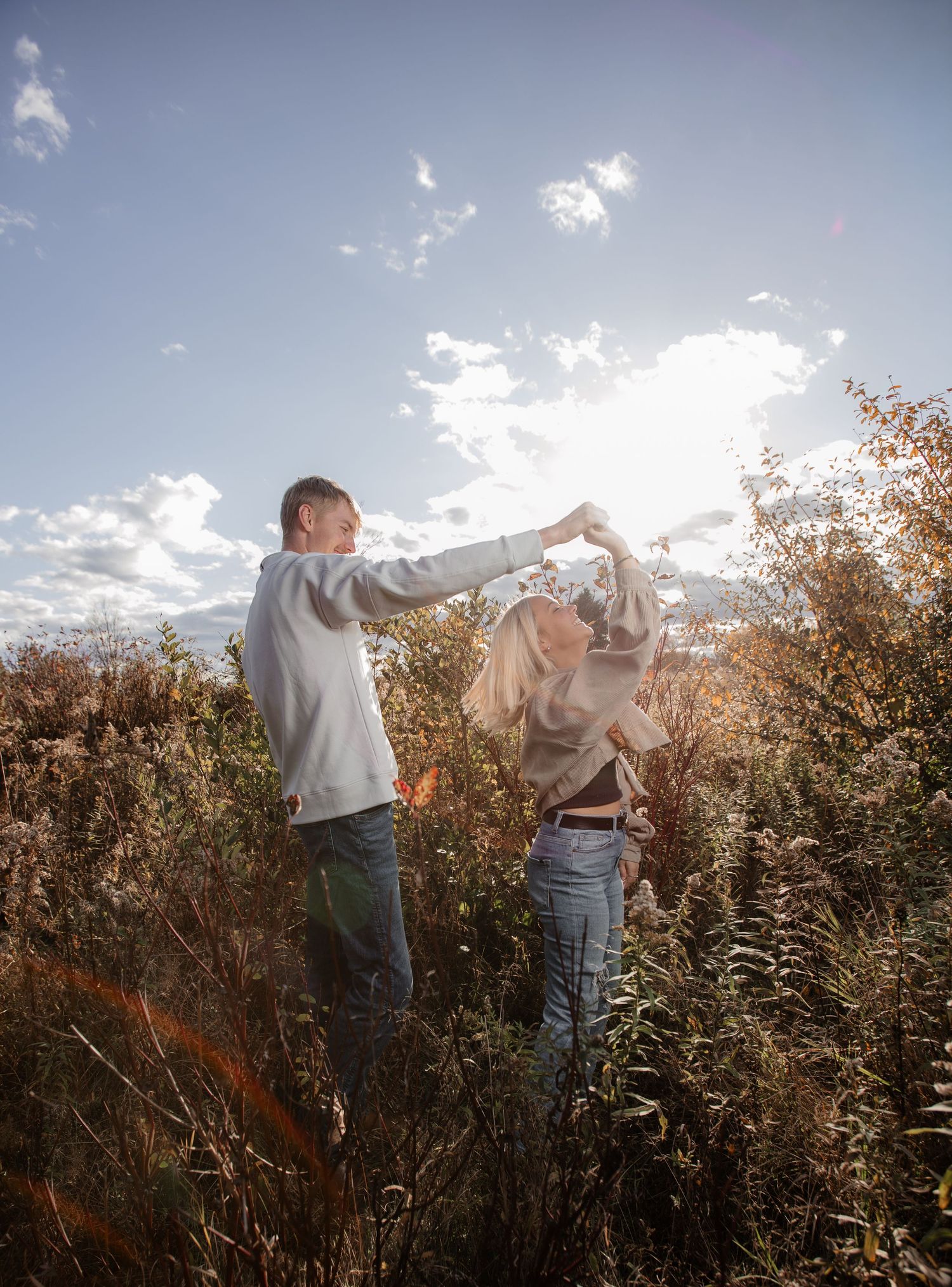 Two people dance playfully in a golden field at sunset with wispy clouds in the bright blue sky.