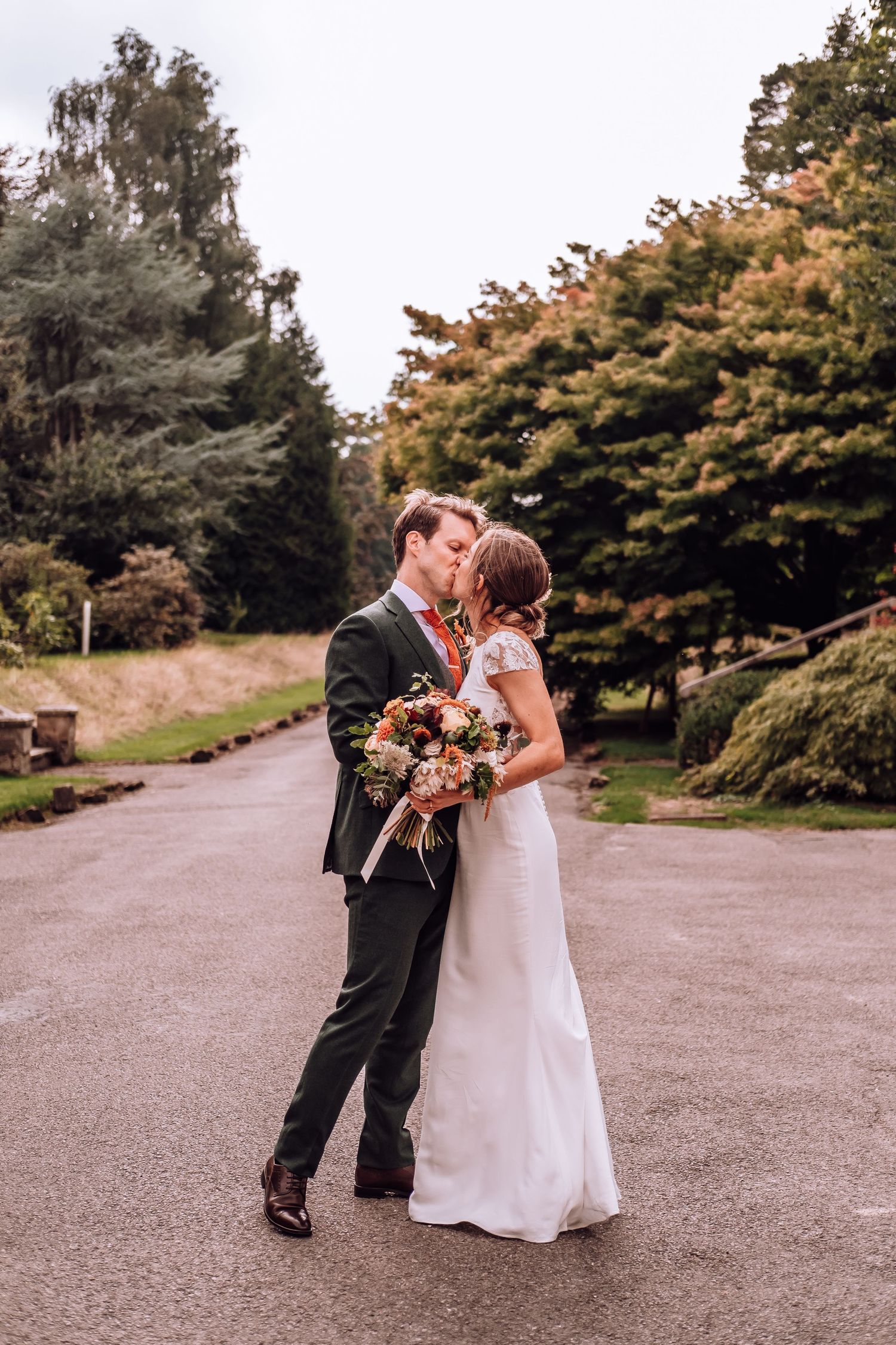 A romantic couple shares an intimate moment on a paved garden path surrounded by autumn trees.
