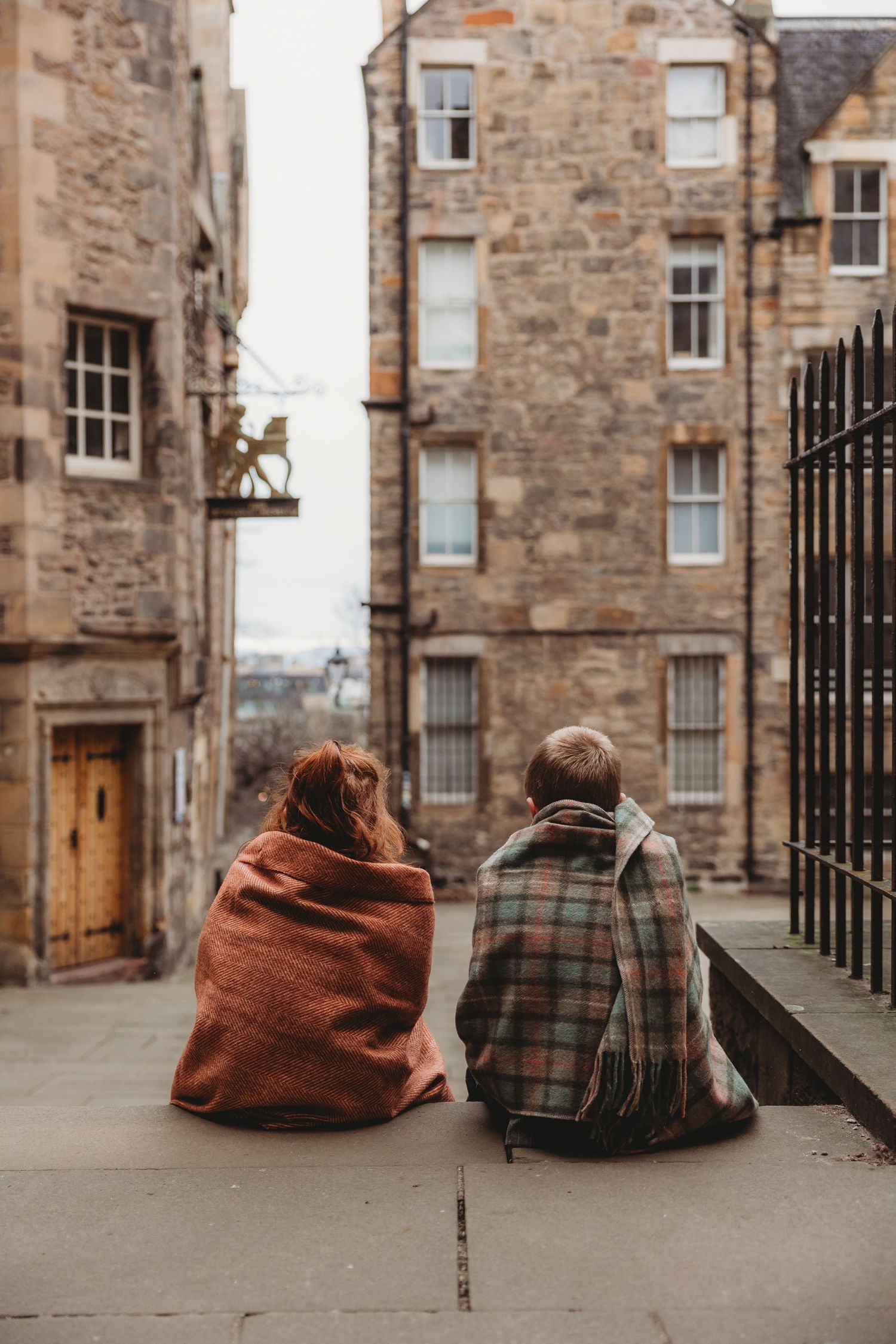 Two people sit together on stone steps in Edinburgh's historic old town wearing plaid and rust colored outerwear.