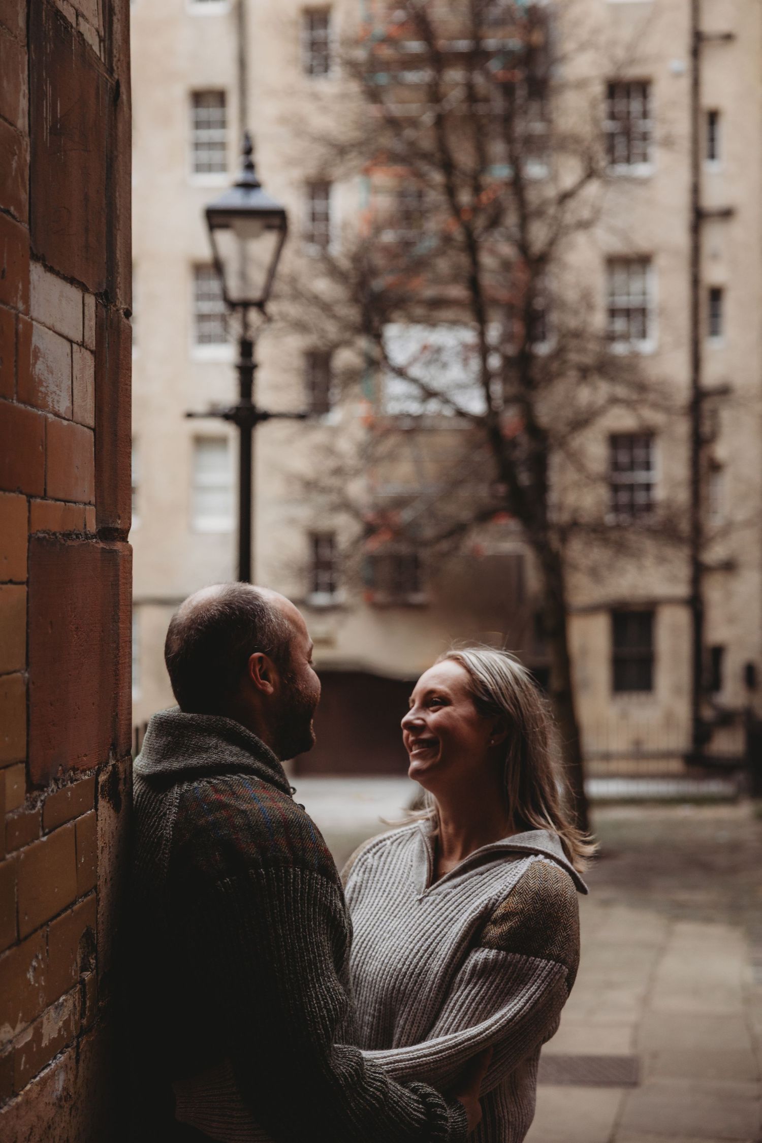 A couple in cozy sweaters share a tender moment while standing against a brick wall on an urban street.