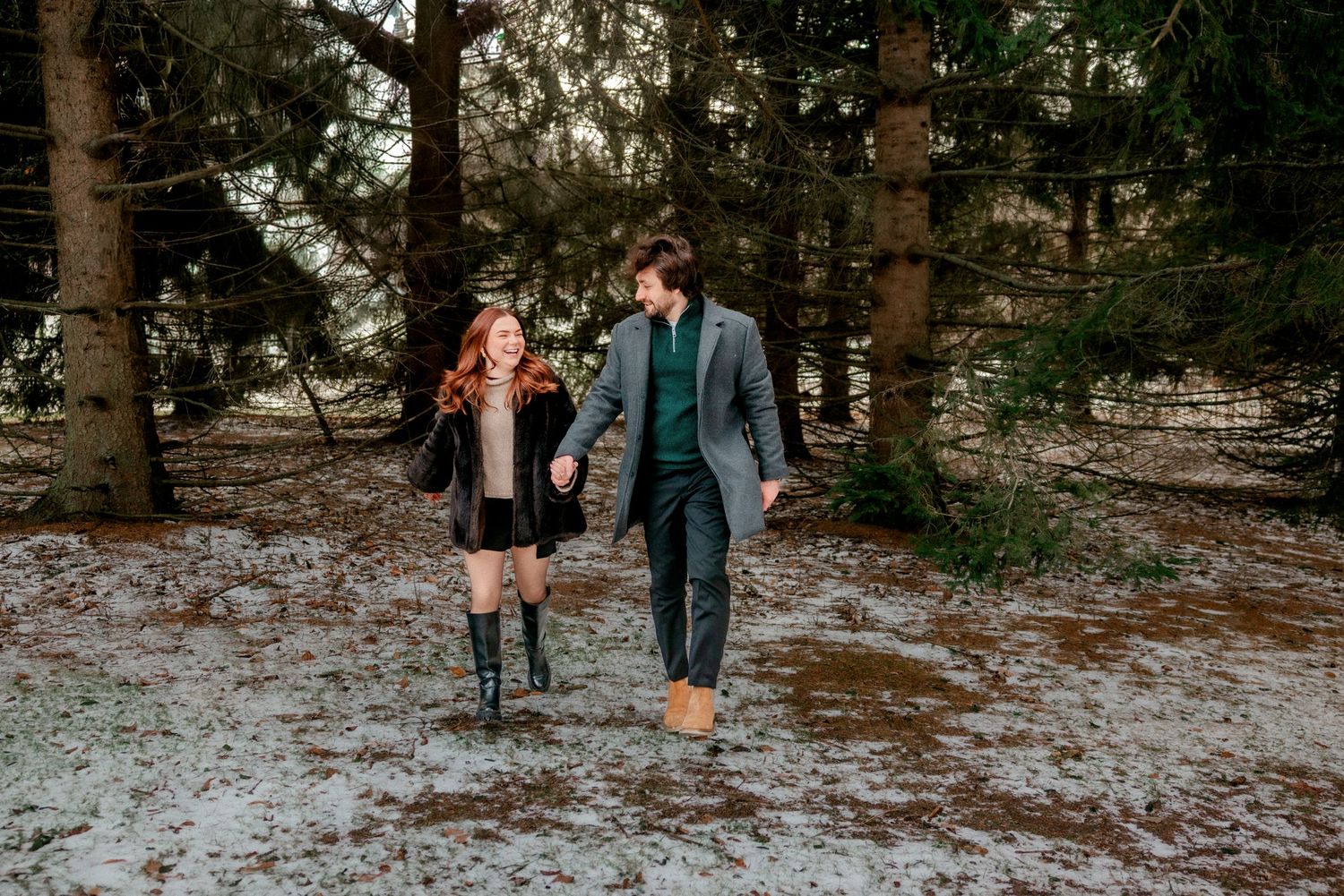 A couple walks together through a snowy forest path during a winter engagement photo session.
