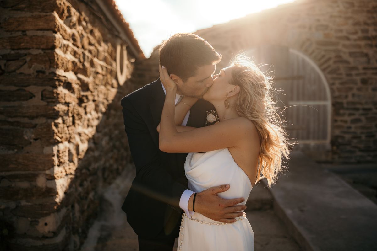 Romantic wedding couple embraces in warm sunset light against rustic stone wall backdrop.