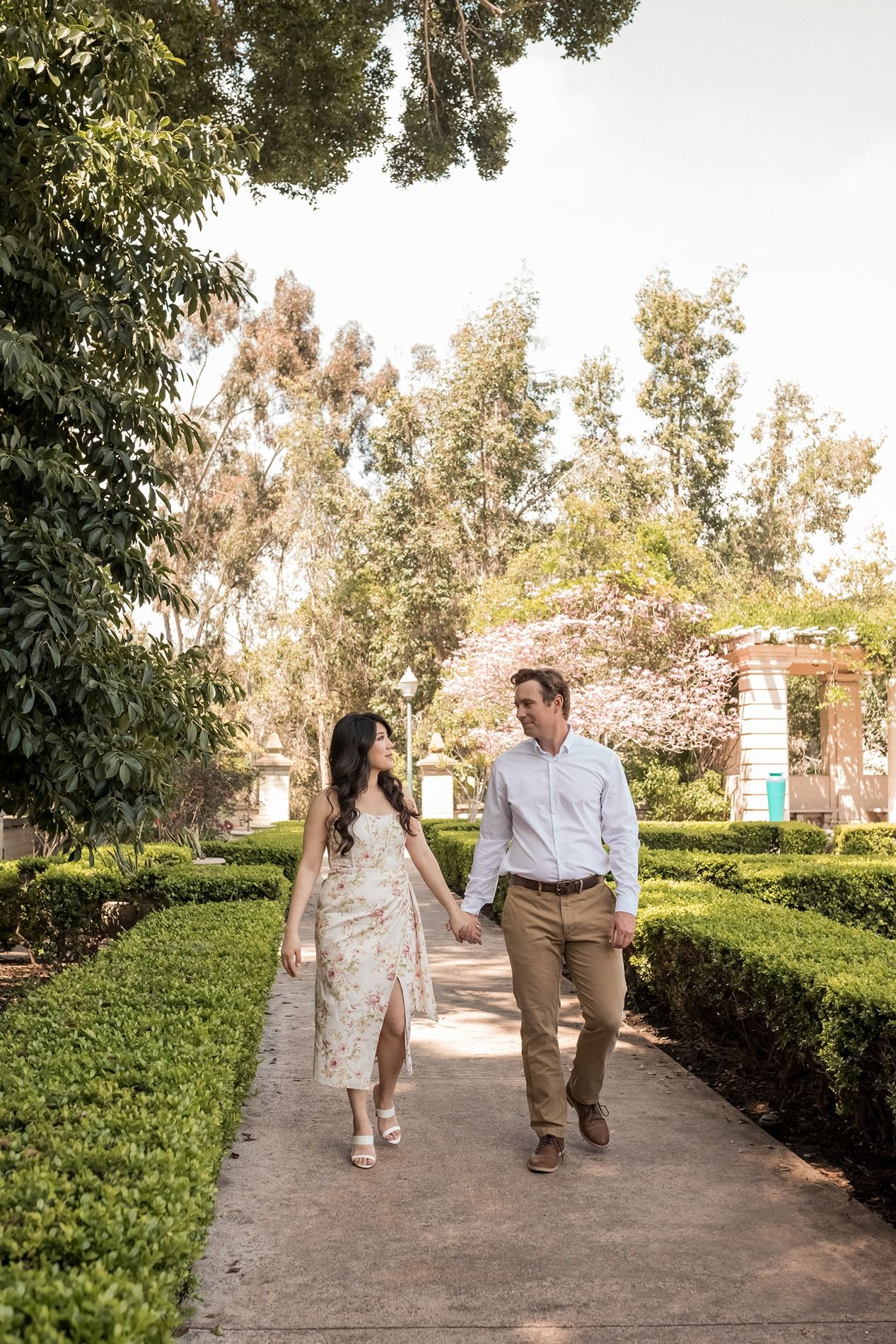 A couple walks together on a garden path lined with manicured hedges and trees on a sunny afternoon at Balboa Park in San Diego.
