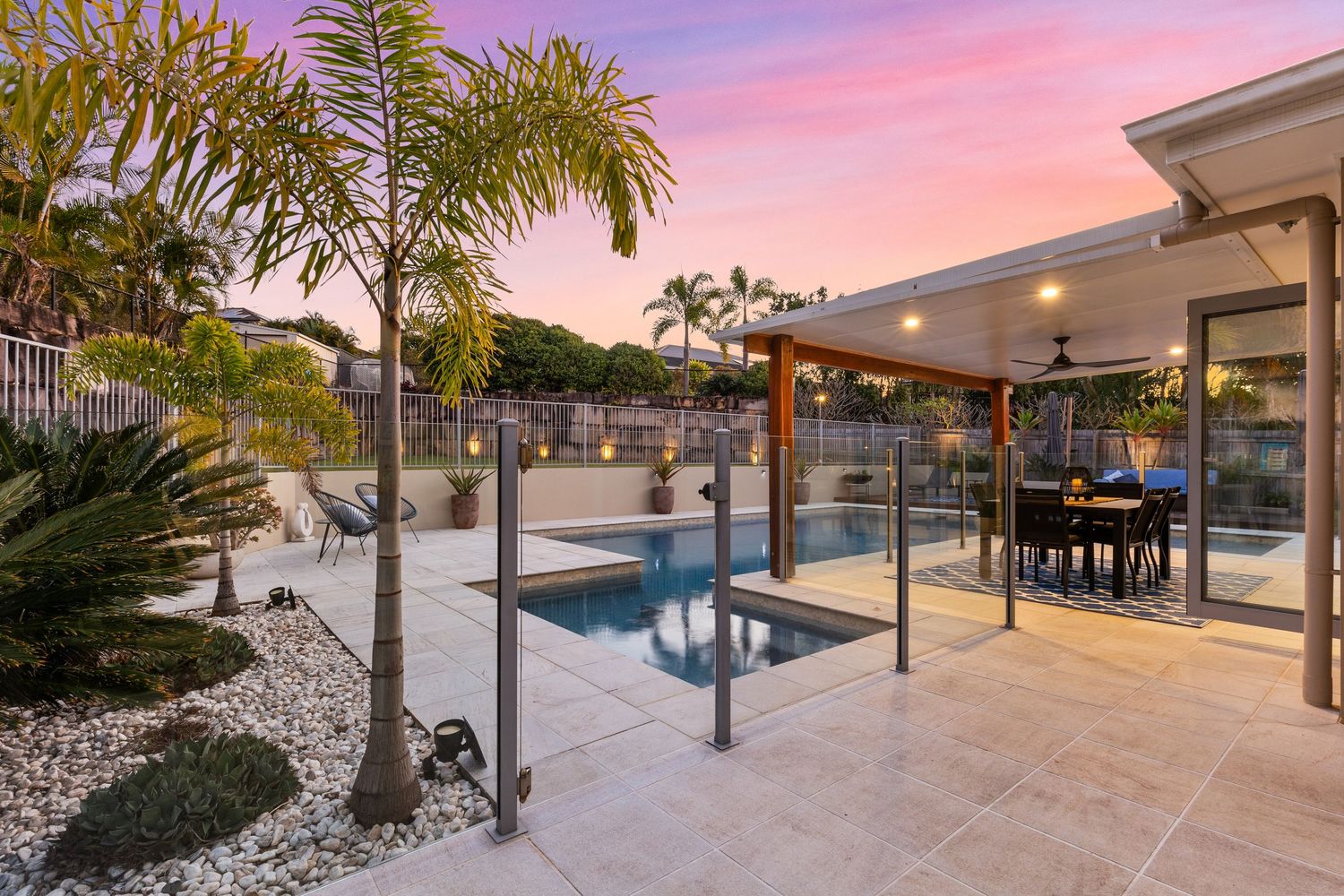 Resort-style backyard with plunge pool, glass fencing, covered alfresco dining, and tropical palms at sunset.