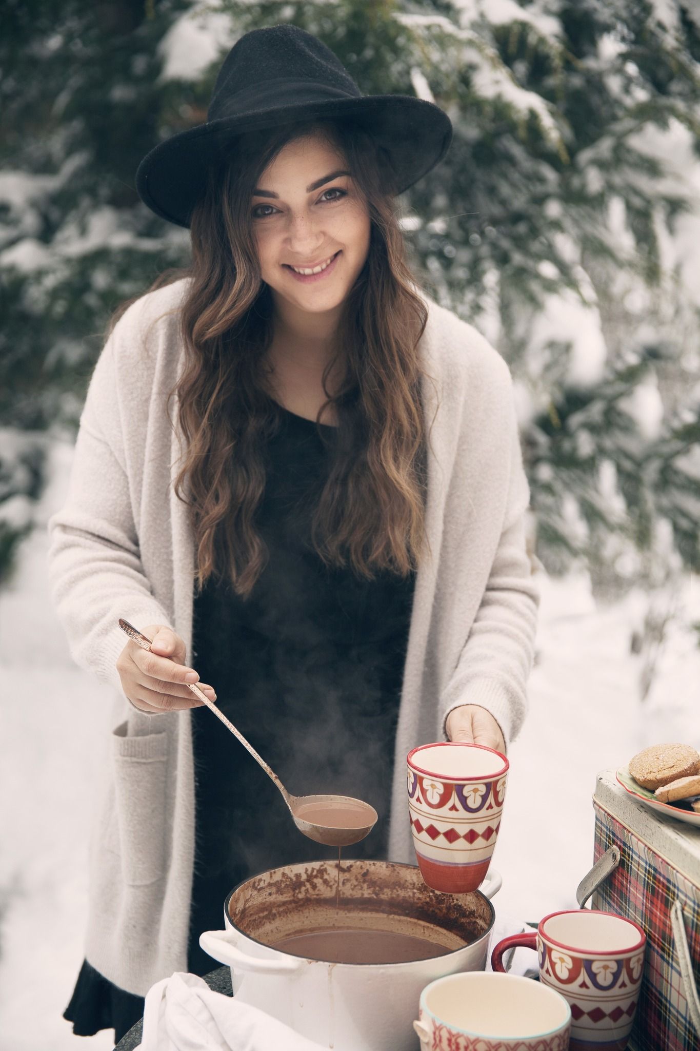 Cozy winter scene with hot chocolate and warm sweaters in a rustic outdoor setting with snowy evergreen branches.