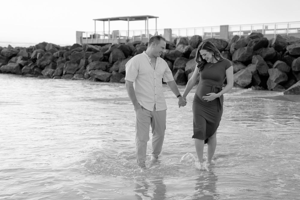 Two people walk hand in hand along a rocky shoreline with waves crashing against the rocks on a sunny day.
