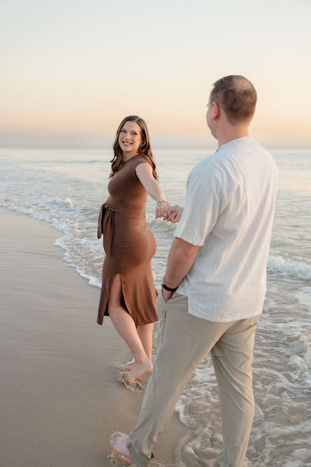 A couple walks hand in hand along a sandy beach at sunset with gentle waves rolling onto the shore.