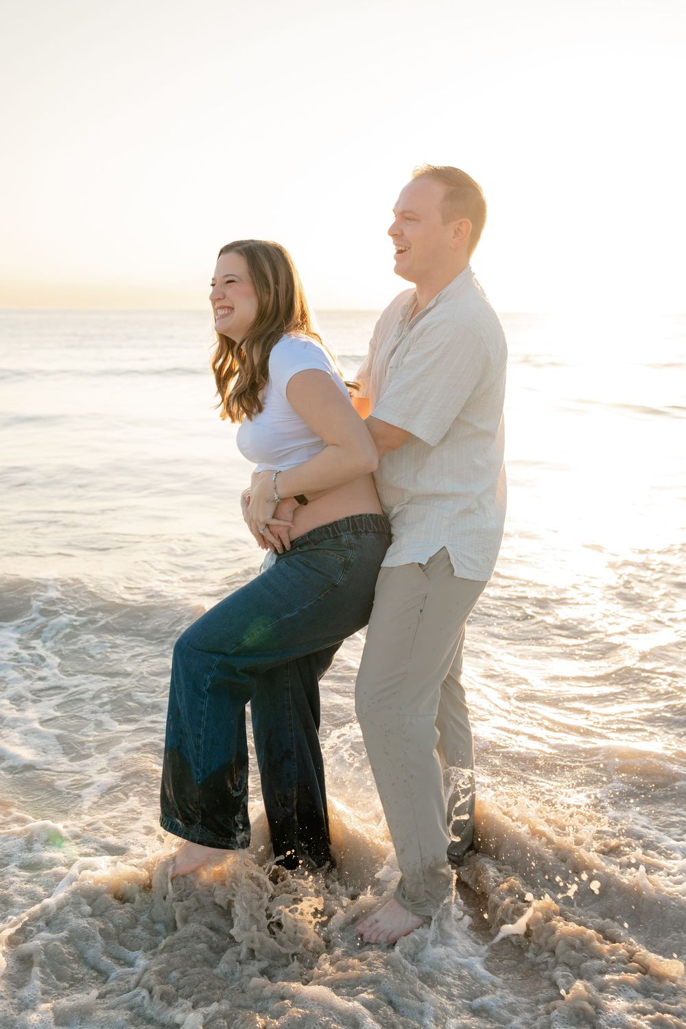 A couple plays and laughs together in the ocean waves at sunset on a sandy beach.