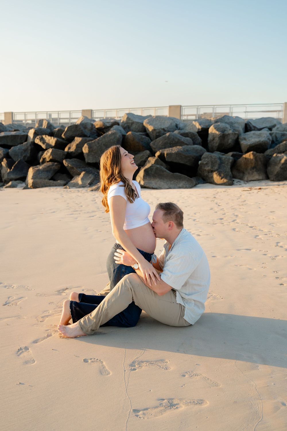 A couple sits together on a sandy beach with dark rocks in the background during a romantic photoshoot.