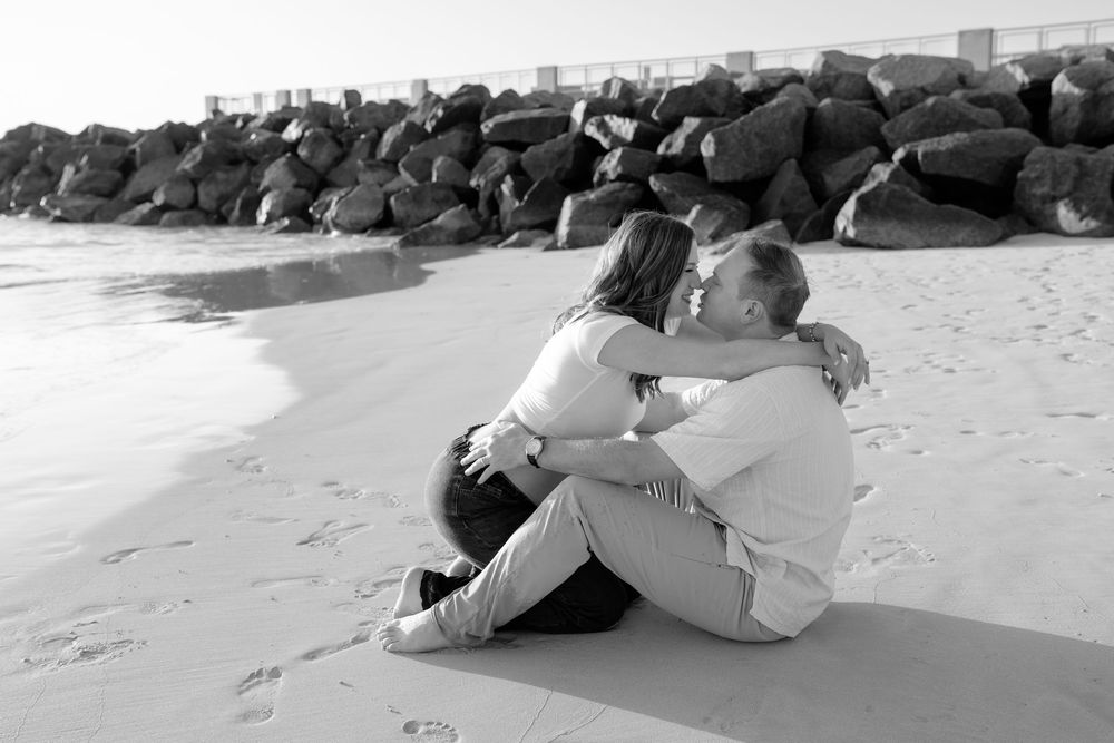 A couple sits close together on the beach near a rocky jetty in black and white.
