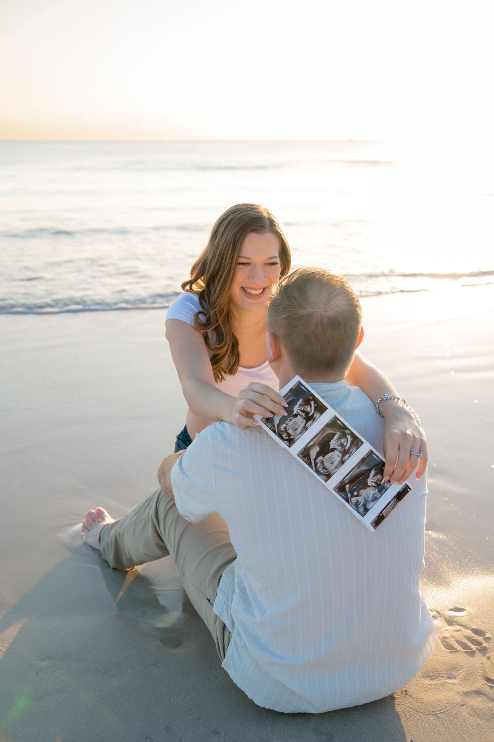 Couple sitting on sandy beach sharing ultrasound photos at sunset.