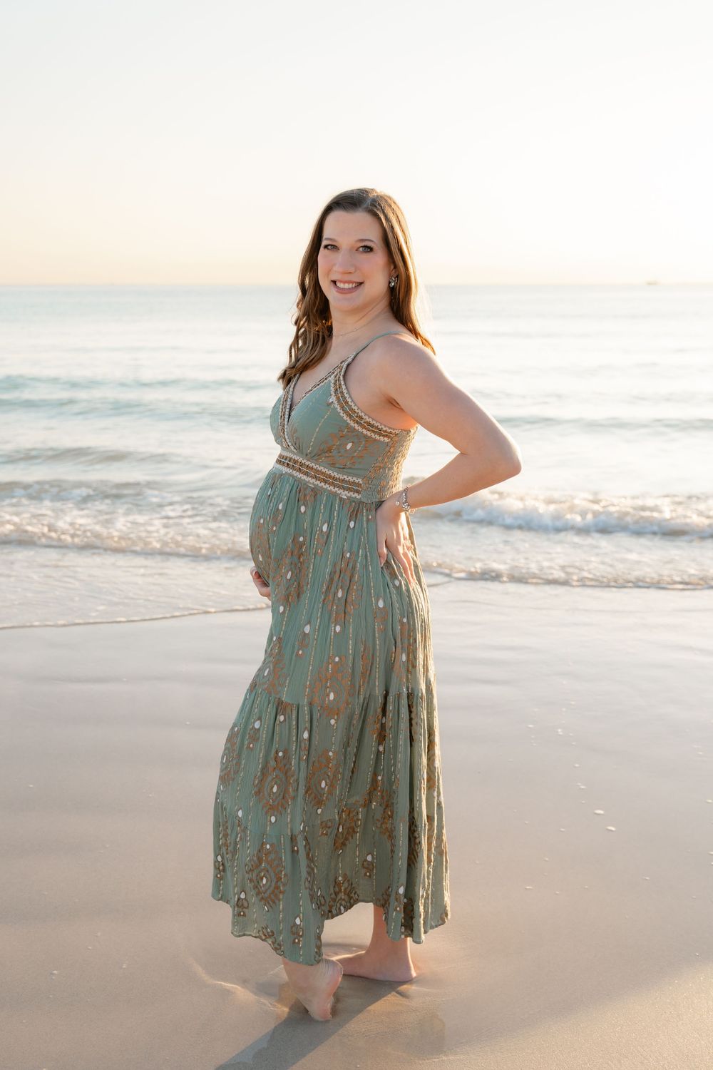 Woman in a flowy sage green dress poses on a sandy beach during golden hour.
