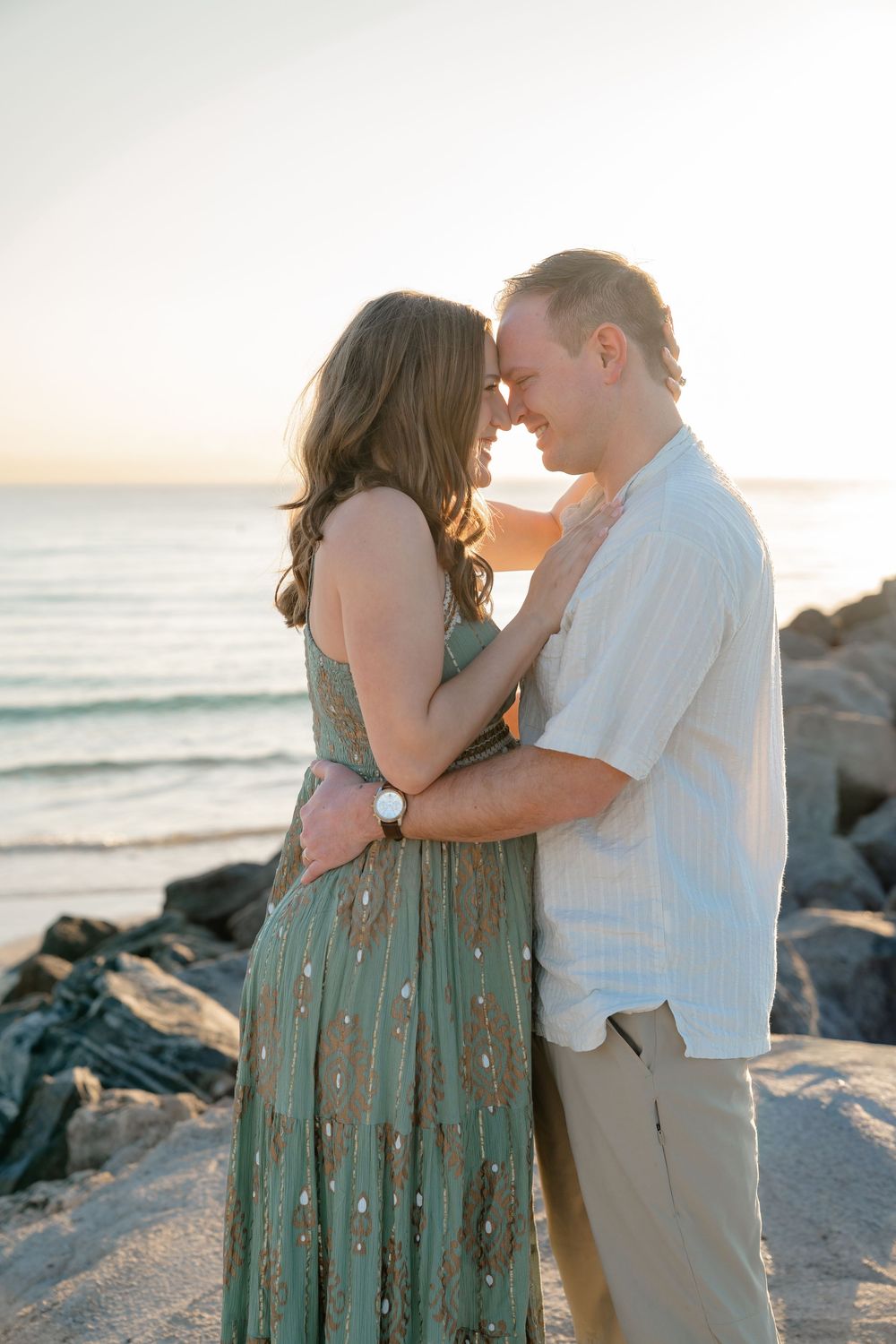 A romantic couple embraces on the beach at golden hour with the ocean and rocks in the background.