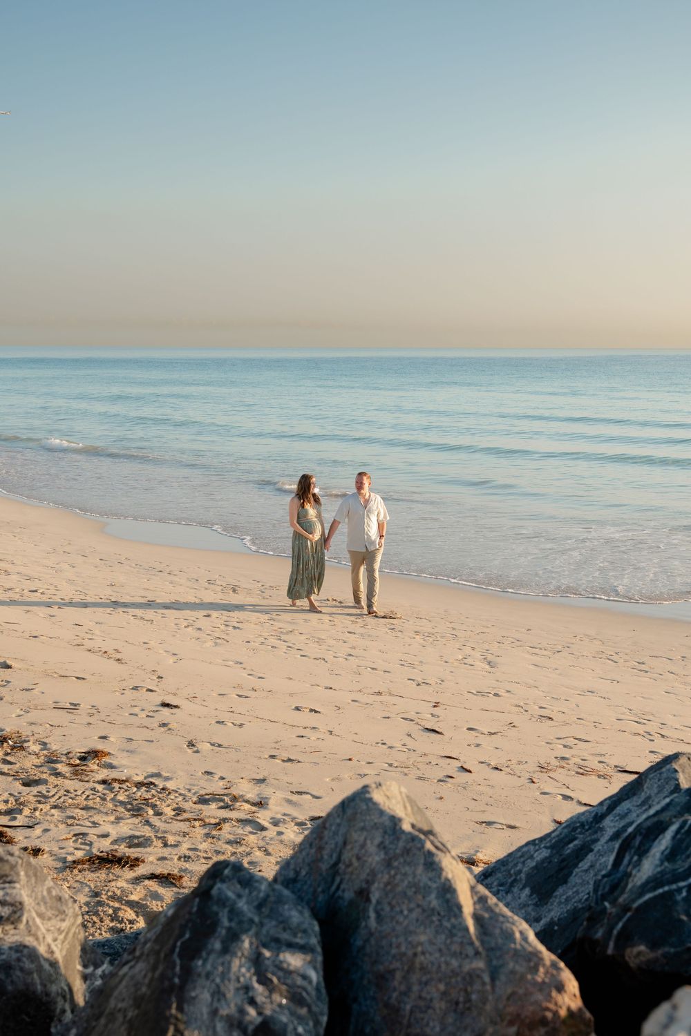 Two figures walk along a serene beach at sunset with dark rocks in the foreground and calm ocean waves in the background.