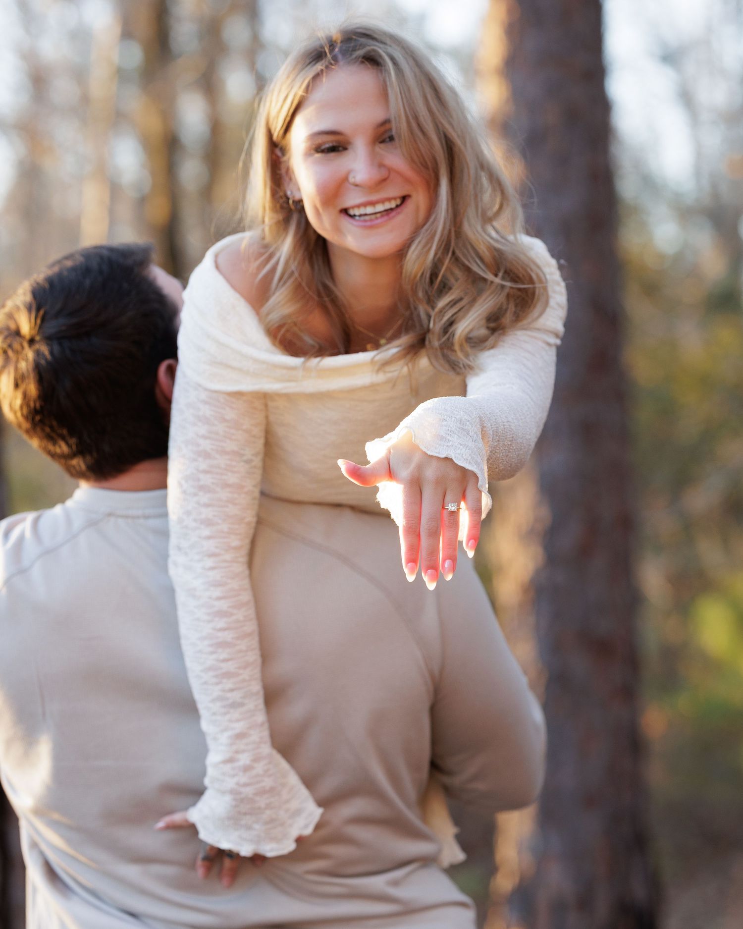 Person in cream sweater displaying engagement ring against autumn forest backdrop.