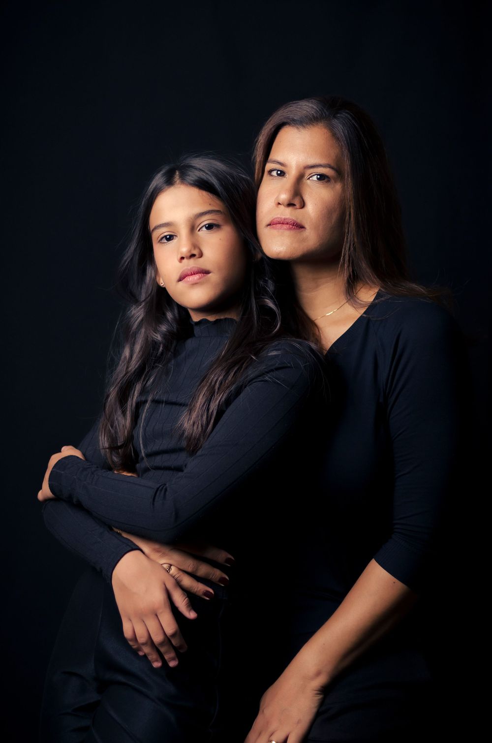 Fotografia de família em estúdio de mãe e filha, abraçadas, com roupas pretas e fundo escuro. Ensaio mãe e filha profissional.
