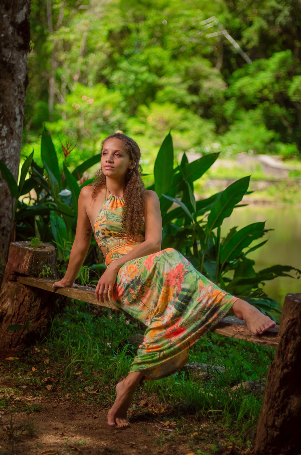 Ensaio feminino na natureza de mulher com vestido longo estampado em tronco de árvore, com paisagem verde e lago. Fotografia ao ar livre.