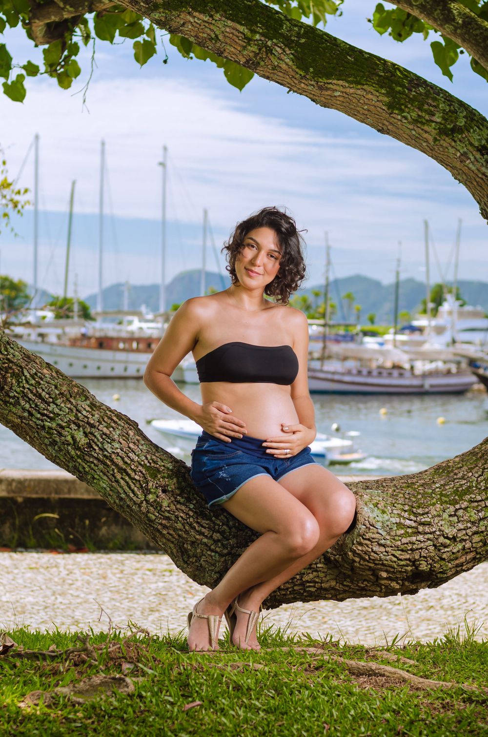 Ensaio de gestante na natureza. Mulher grávida sentada em árvore, de top e shorts jeans, com fundo de barcos. Fotografia de maternidade.