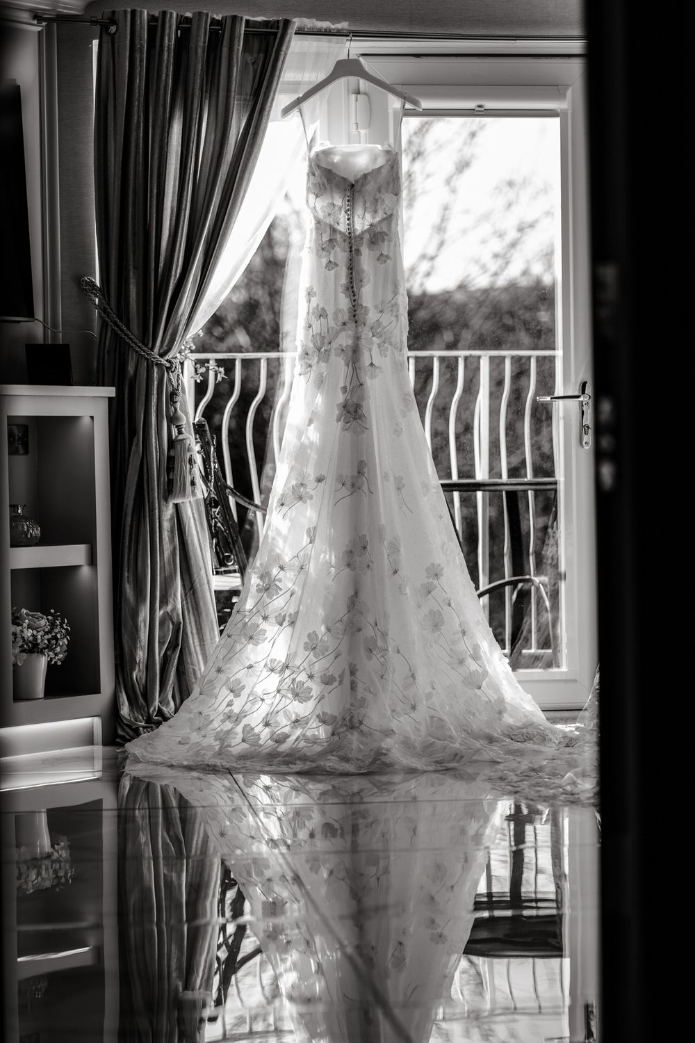 A wedding dress hangs in front of a window creating a silhouette effect in black and white photography.