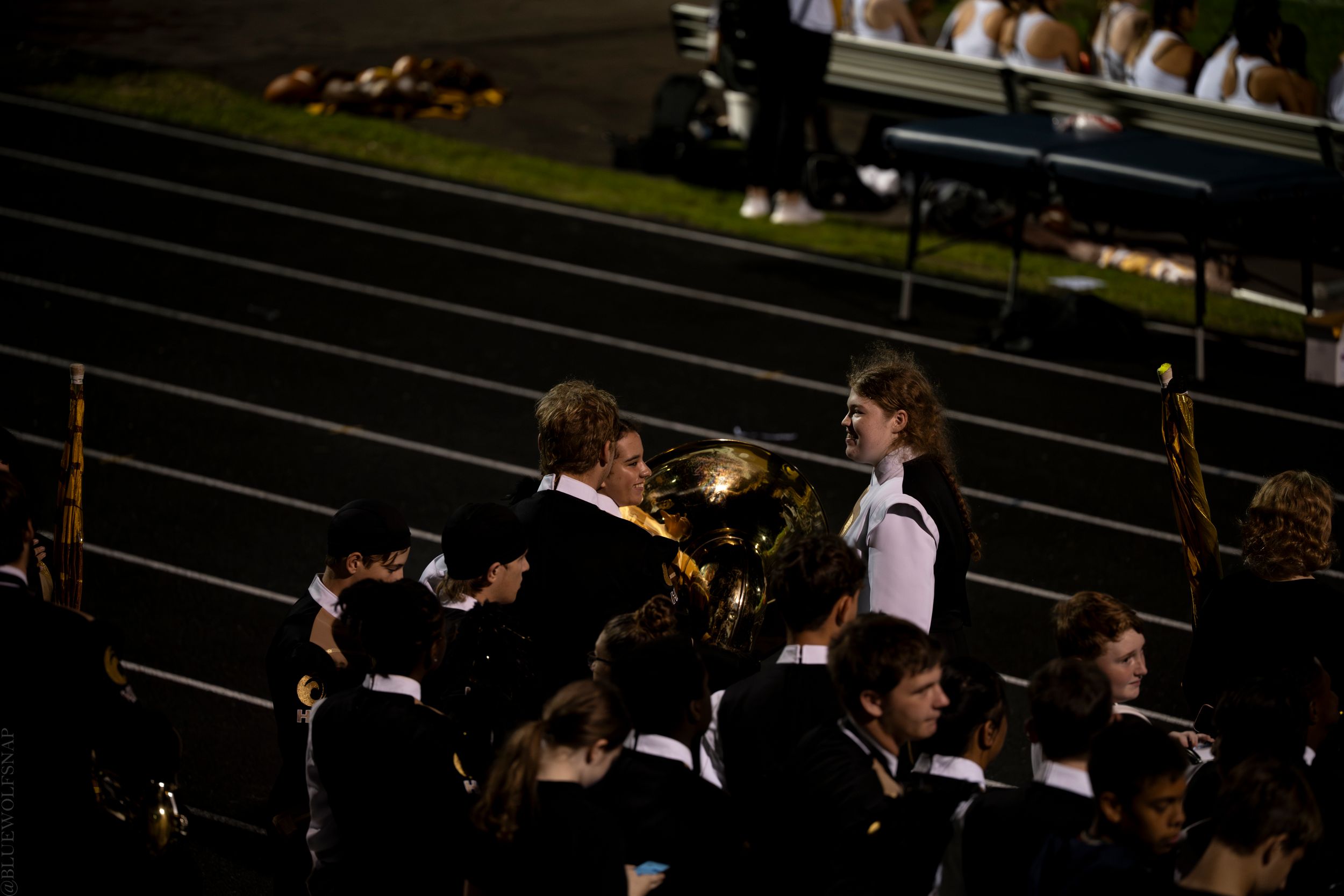 Marching band members perform at night, with a tuba player in focus during an outdoor stadium event.