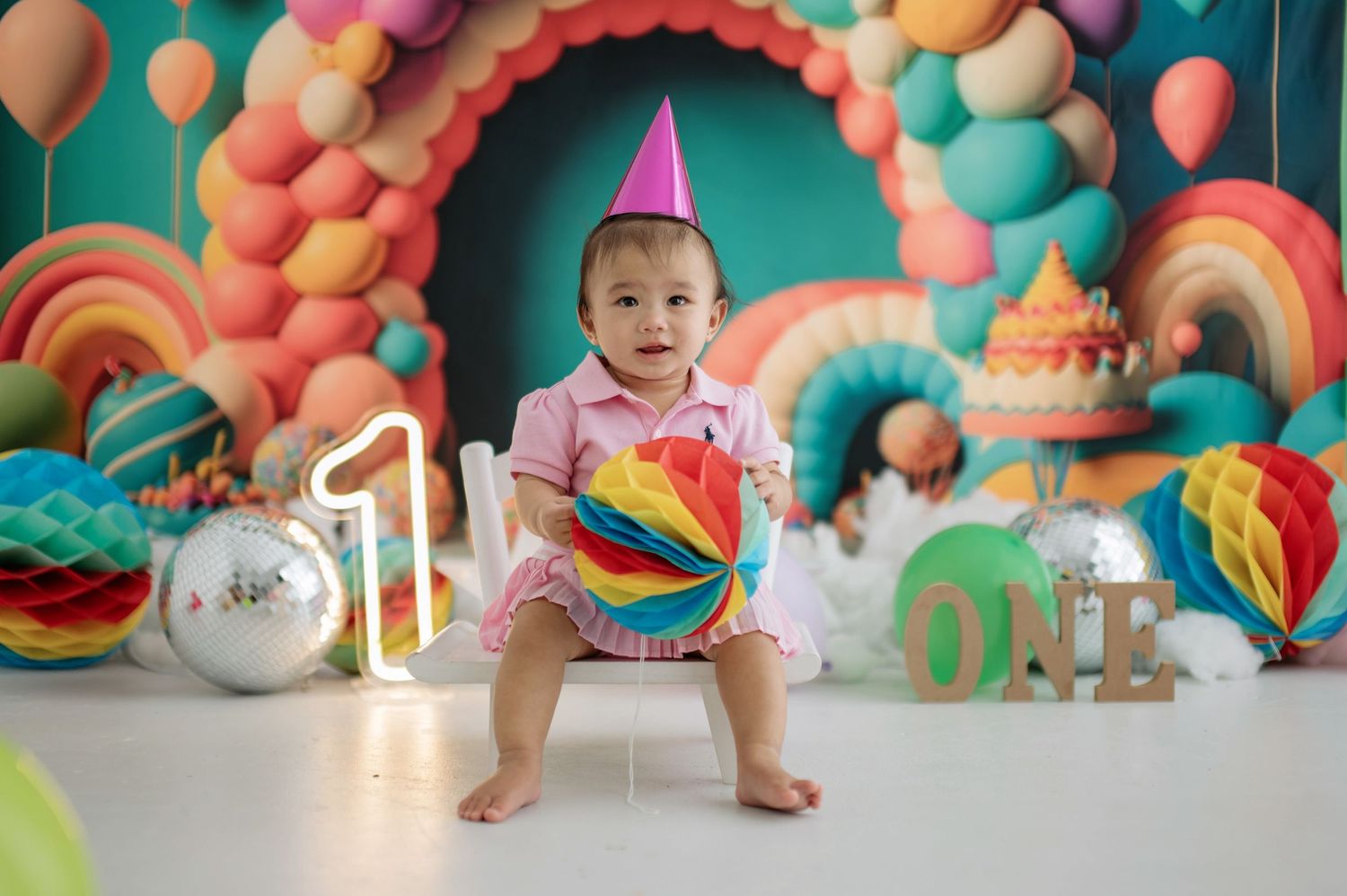 Colorful first birthday party setup with rainbow balloon arch, decorative swirls and party hat against turquoise backdrop.