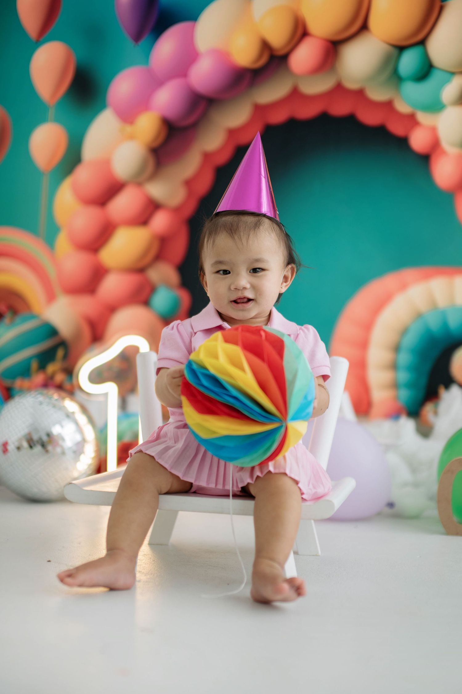Colorful balloon arch decorates a festive birthday setup with teal background and pink party accessories.