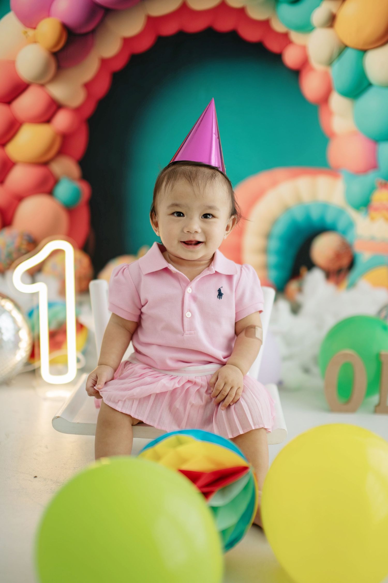 First birthday party setup with colorful balloon arch, neon number one sign, and festive party decorations in pink and teal.