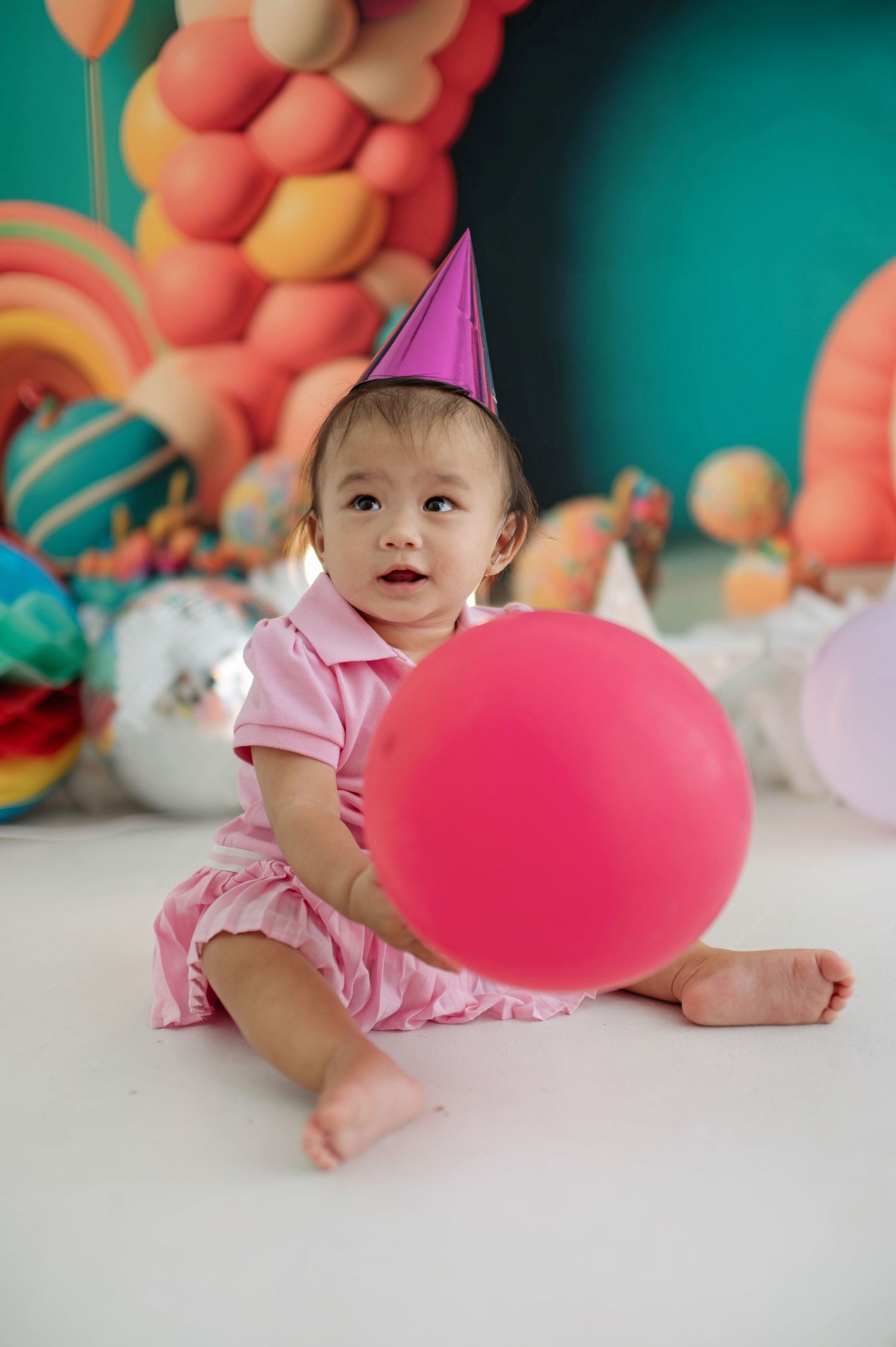 A baby in pink attire and party hat sits on white floor with colorful balloon decorations in background.