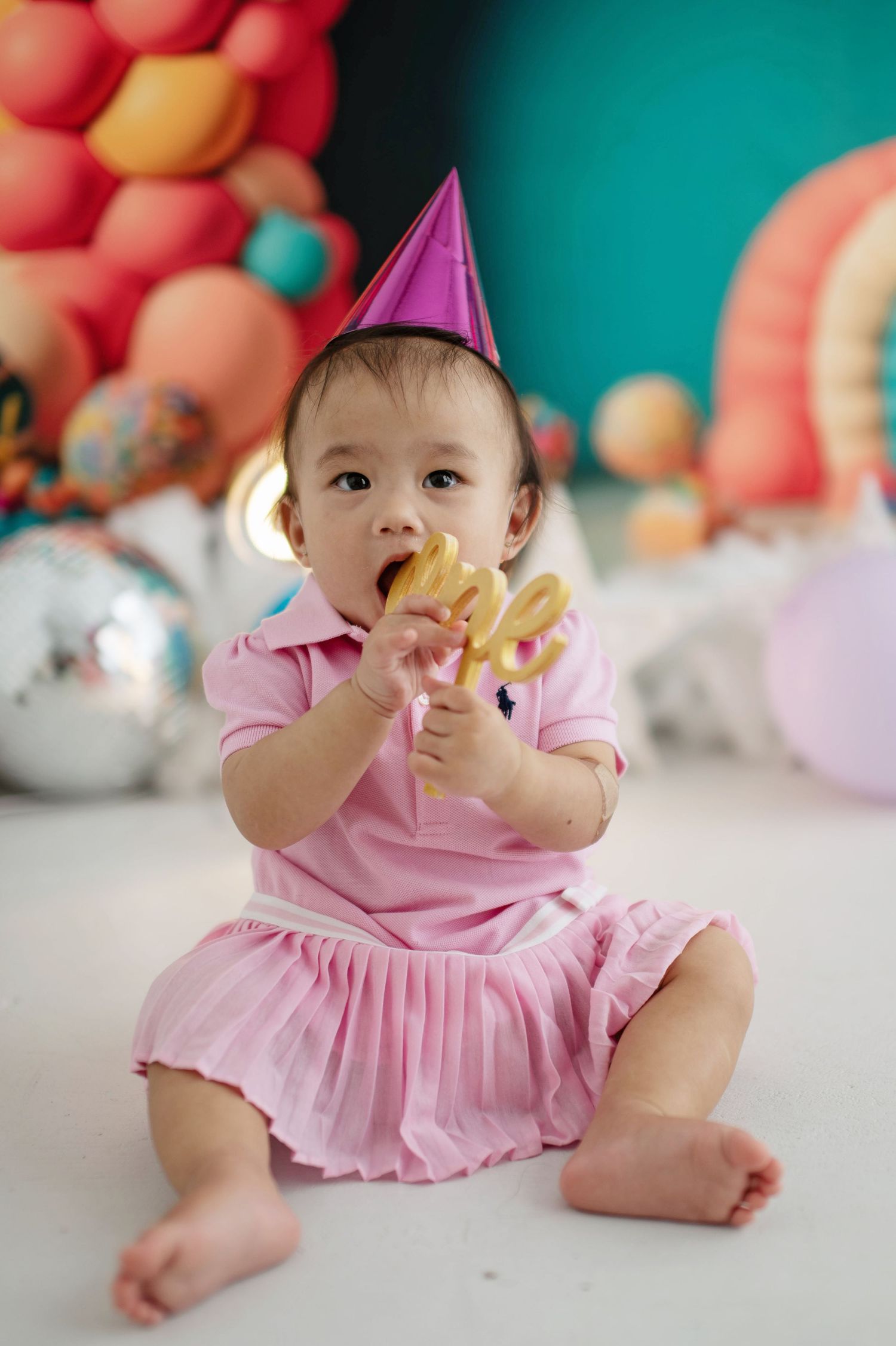 Baby wearing pink ruffled dress sits on floor with colorful balloon decorations in background during birthday celebration.