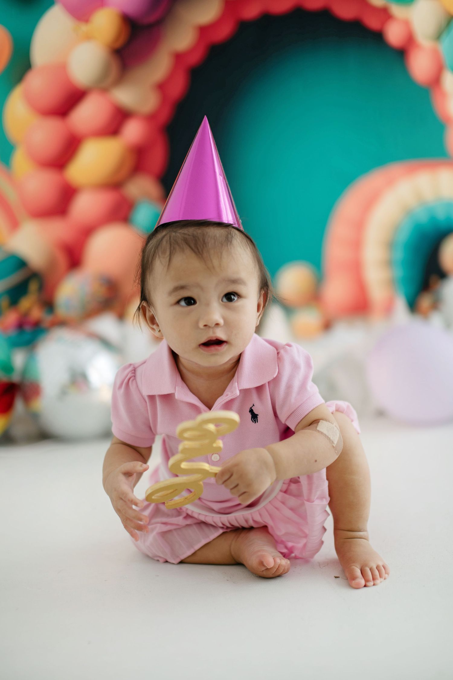 A baby in a pink outfit and party hat lies on the floor with colorful balloon decorations in the background.
