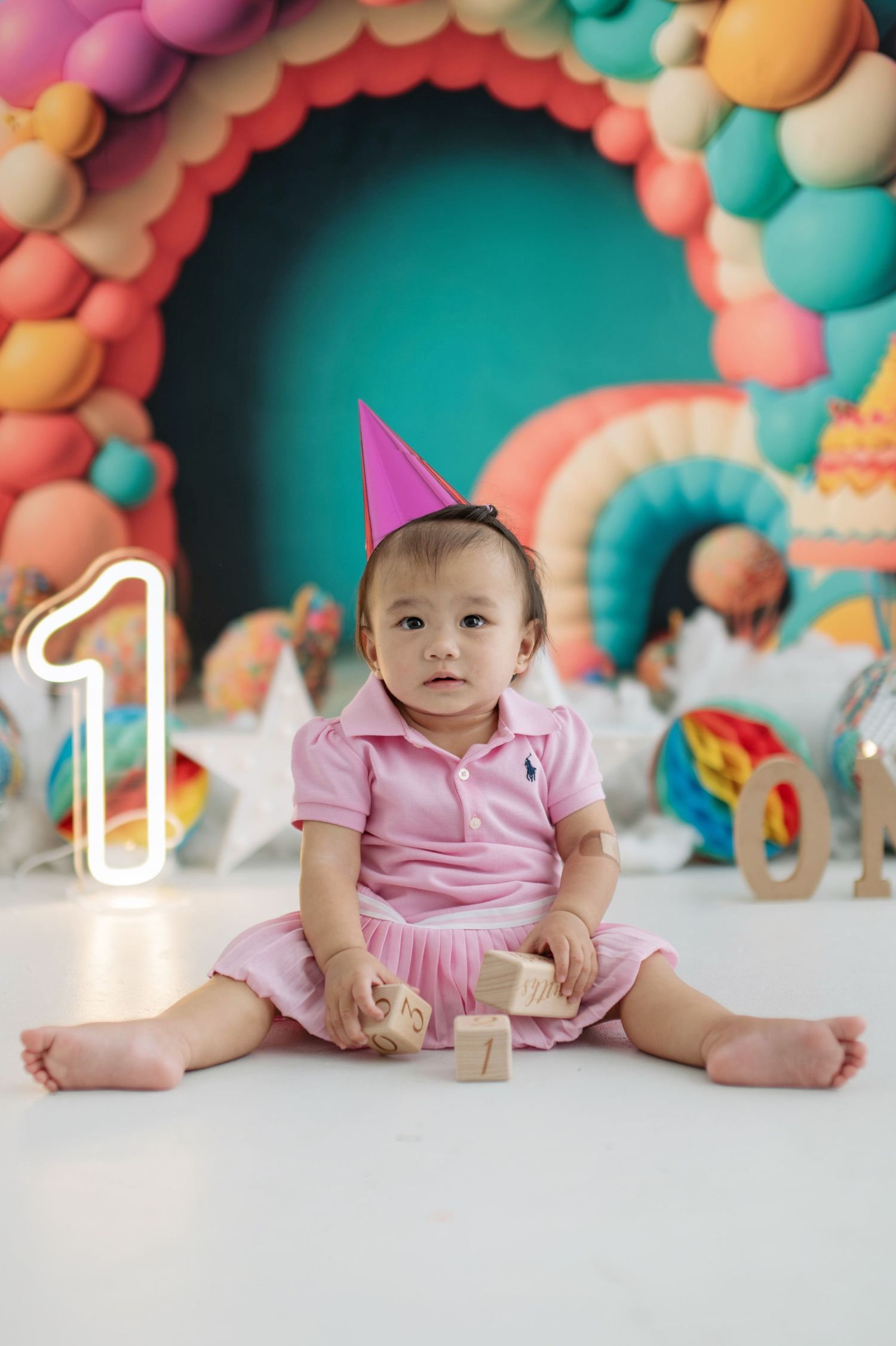 First birthday celebration with pink party dress and colorful balloon arch backdrop in pastel colors.
