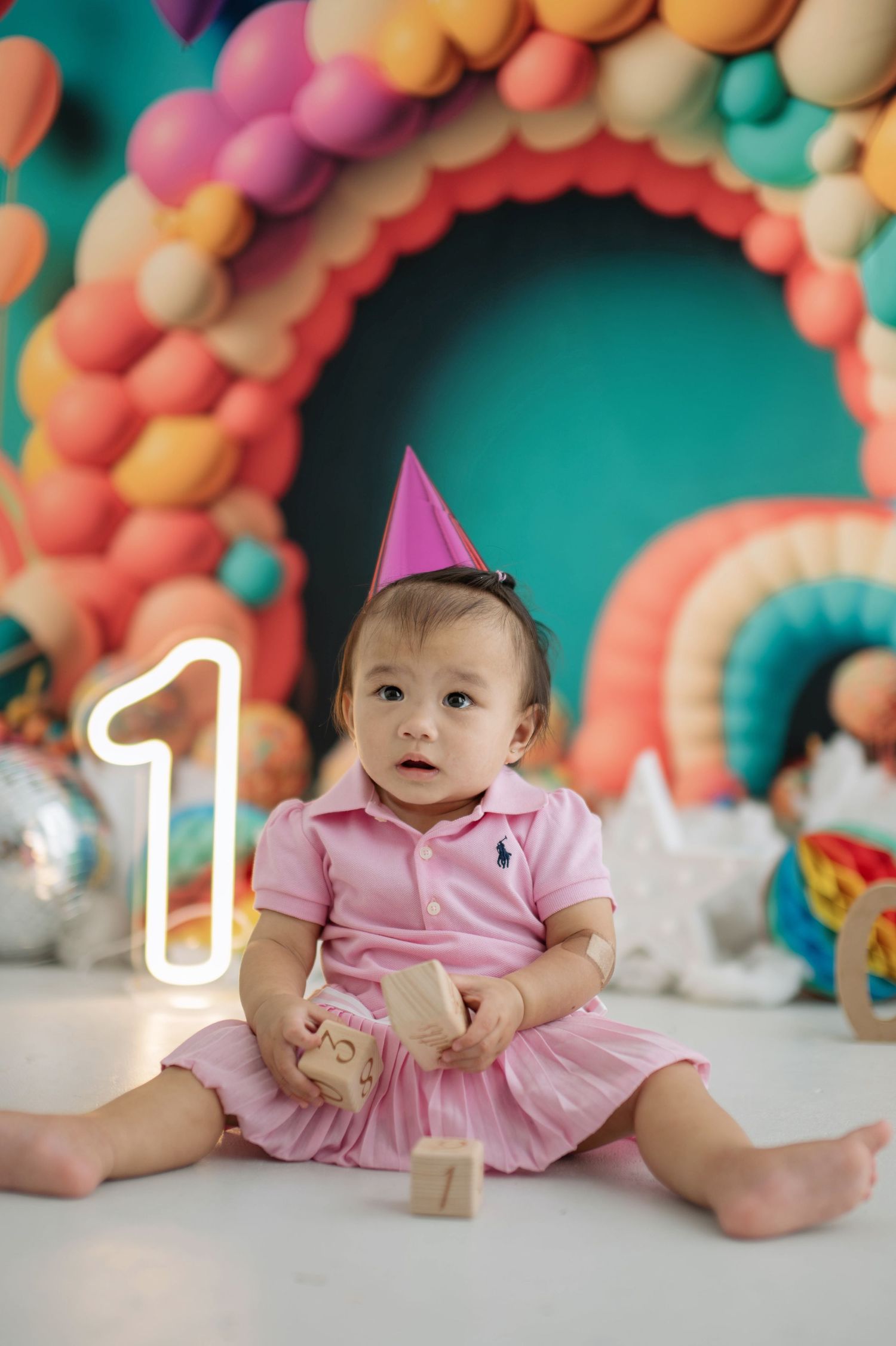 Colorful balloon arch backdrop with a baby in pink dress sitting on the floor during a birthday photoshoot.