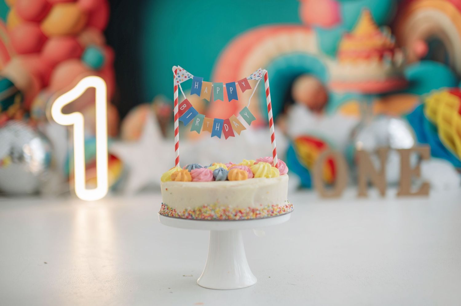 Colorful birthday cake with rainbow sprinkles and candles displayed on a white cake stand against a blurred background.