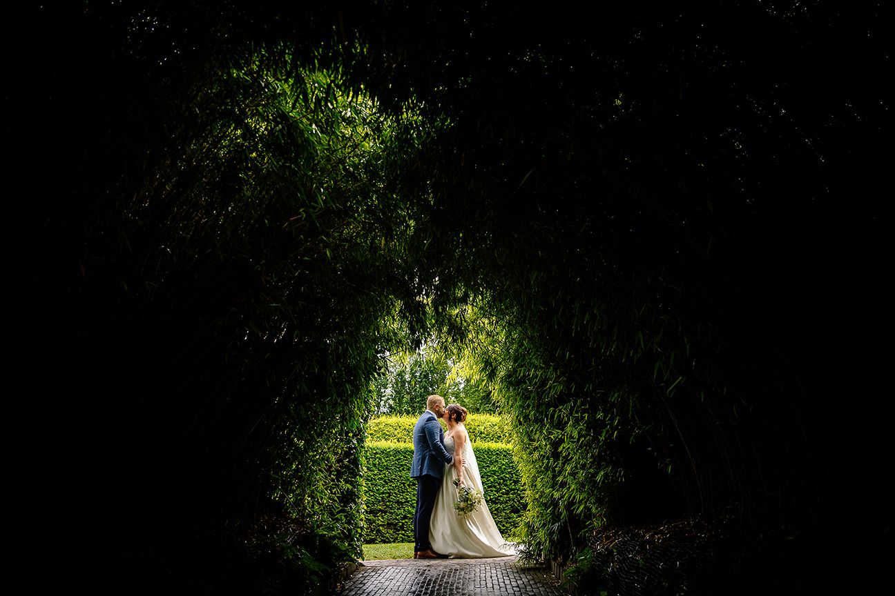 A couple stands in a romantic garden tunnel covered with lush green foliage and natural light filtering through.