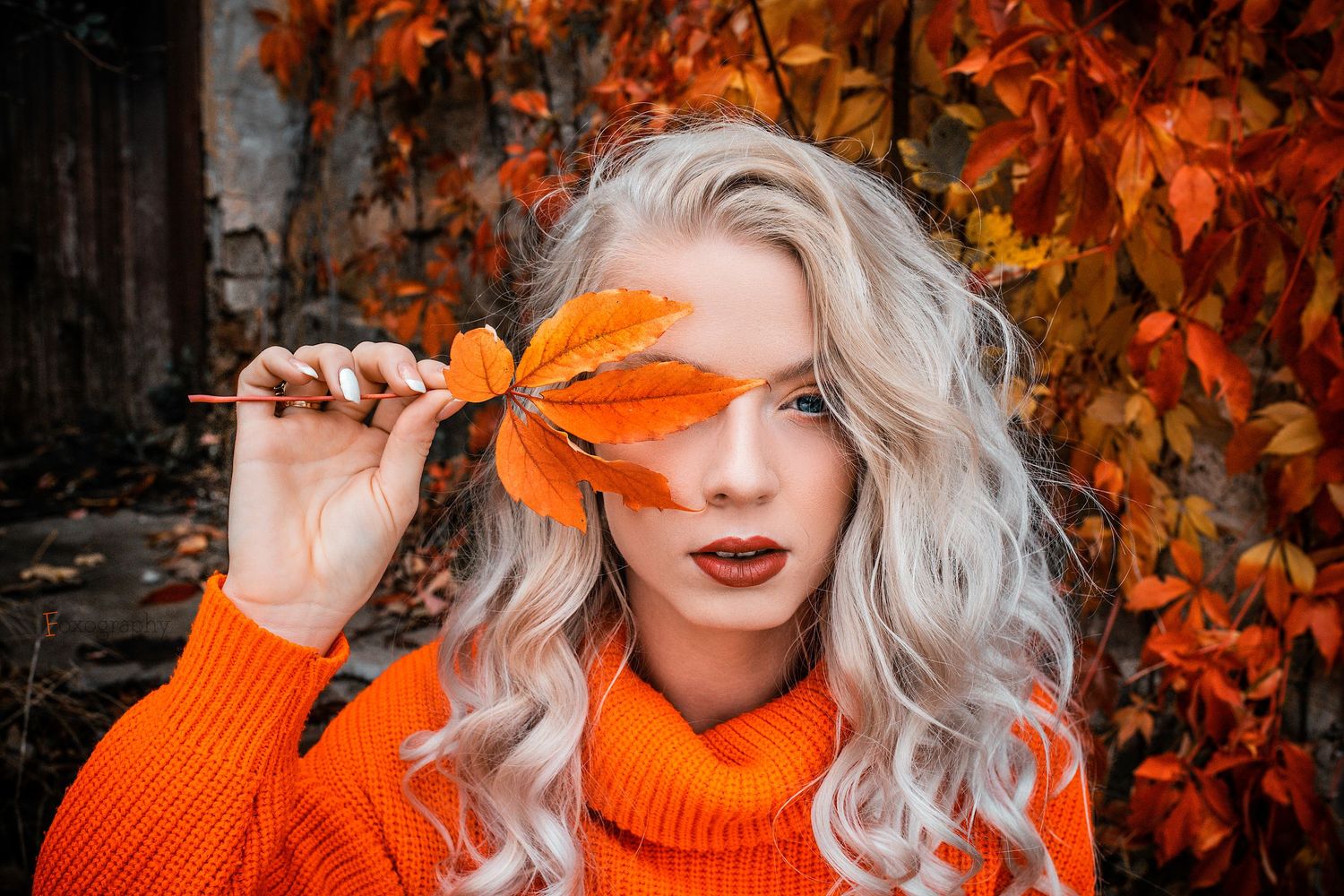 Blondínka v oranžovom svetri si vonku drží jesenný list pred jedným okom. Jesenné fotenie, Žilina.