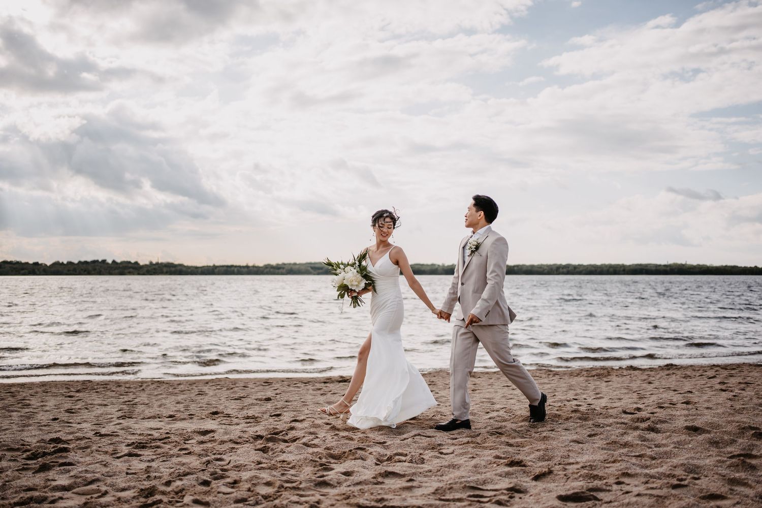 A couple in wedding attire walks hand in hand along a scenic beach under cloudy skies at sunset.