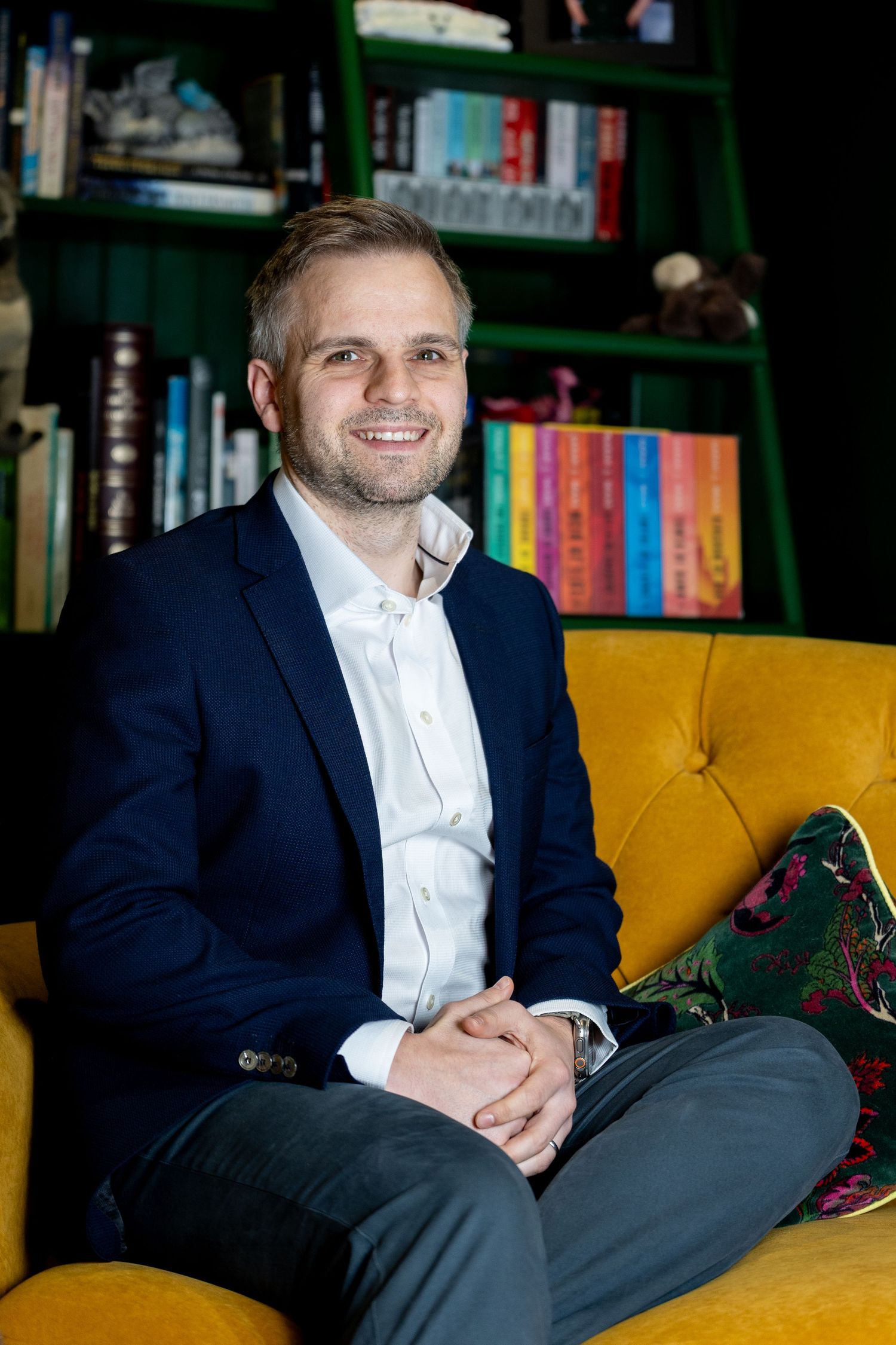 Professional wearing navy suit and white shirt sits on yellow couch in dark moody setting.