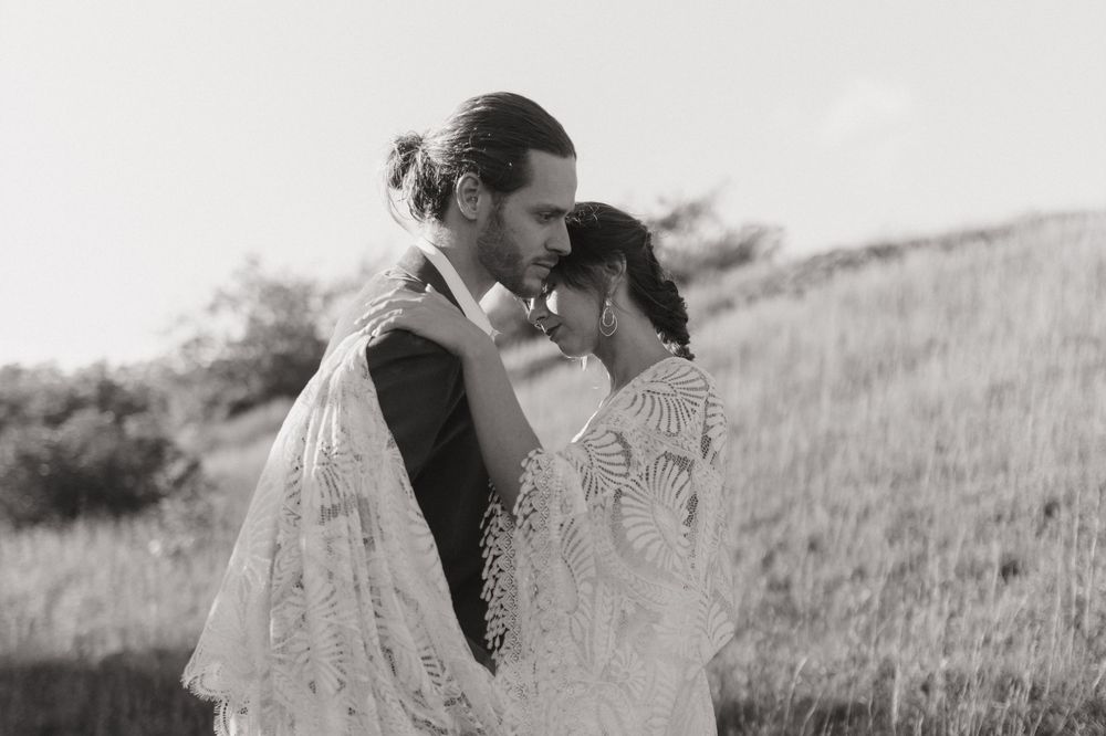 A romantic couple in vintage attire embraces in a sunlit meadow while wearing white lace and formal clothing.
