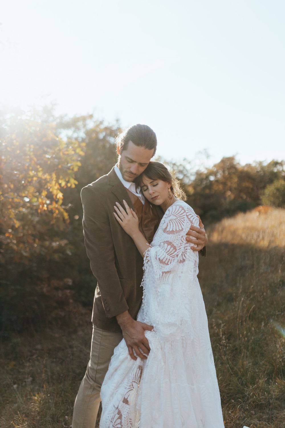 A couple embraces in a romantic sunset moment, with the bride wearing a delicate lace gown against an autumn backdrop.